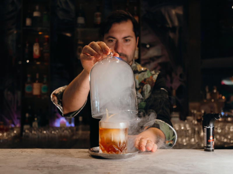 Giacomo Giannotti lifting the glass cover of the Great Gatsy, an Old-Fashioned style cocktail from Paradiso, which uses smoke.